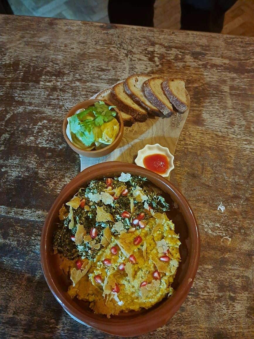 Roast squash in a terracotta dish topped with pomegranate seeds, shaved cheese and herbs, with bread alongside