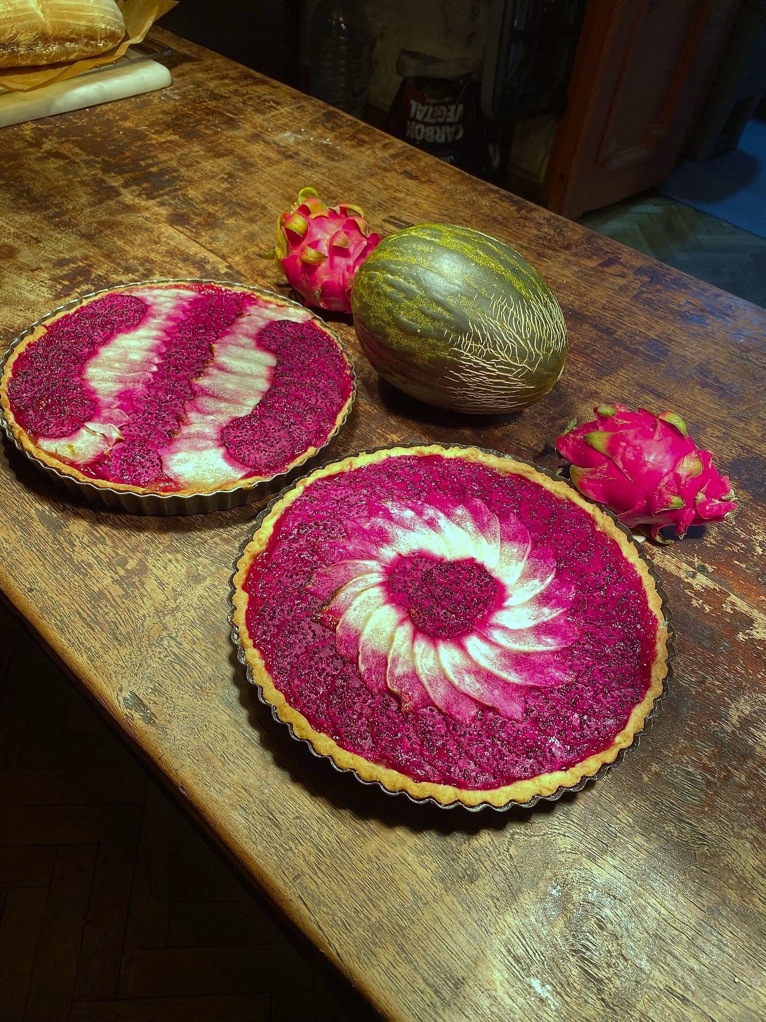 Two tarts topped with pink and white dragon fruit in spiral and striped patterns, with whole fruit on a wooden table