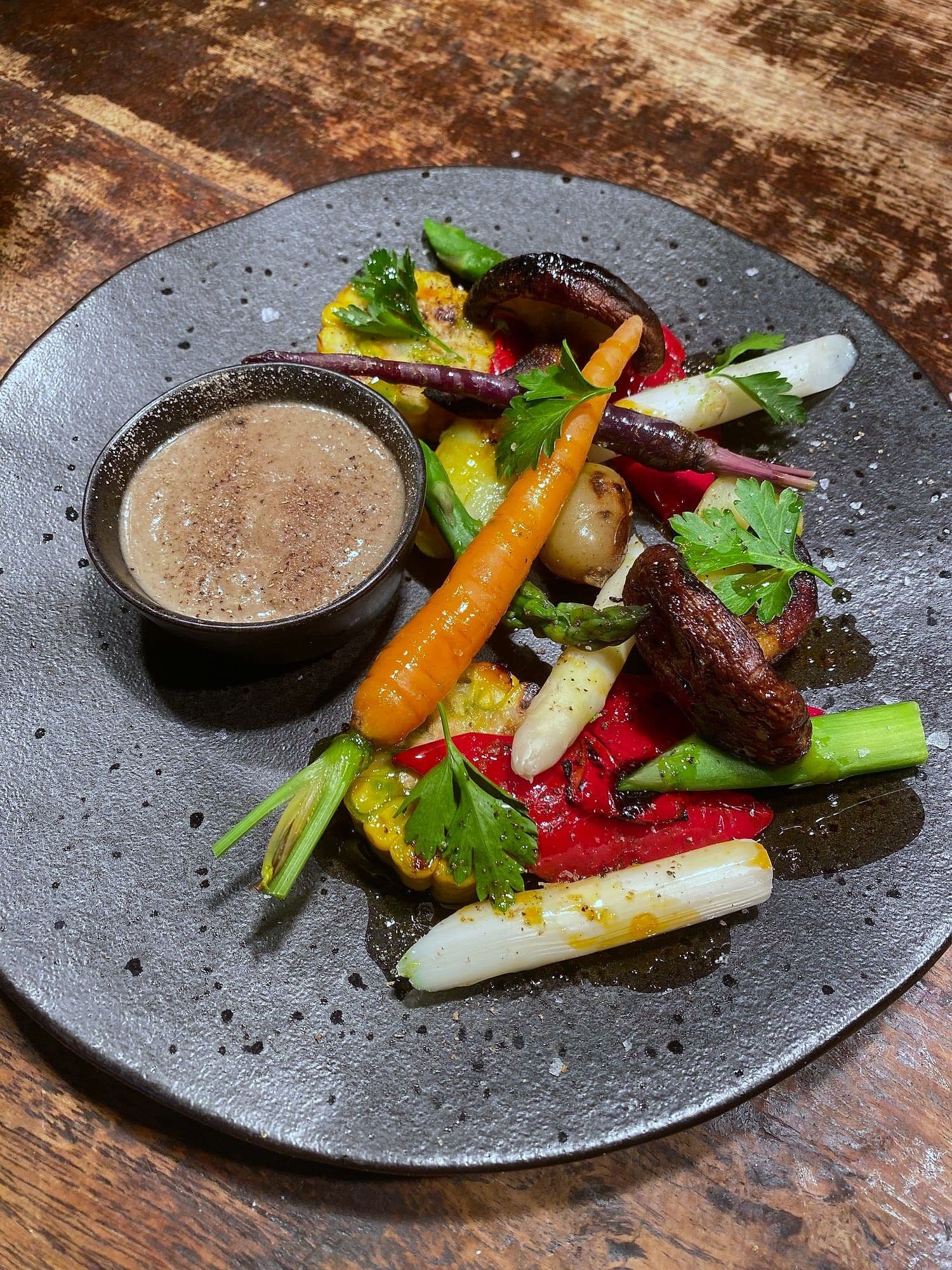 Grilled baby carrots, white asparagus, peppers and corn on a dark plate with a pot of smoked cream