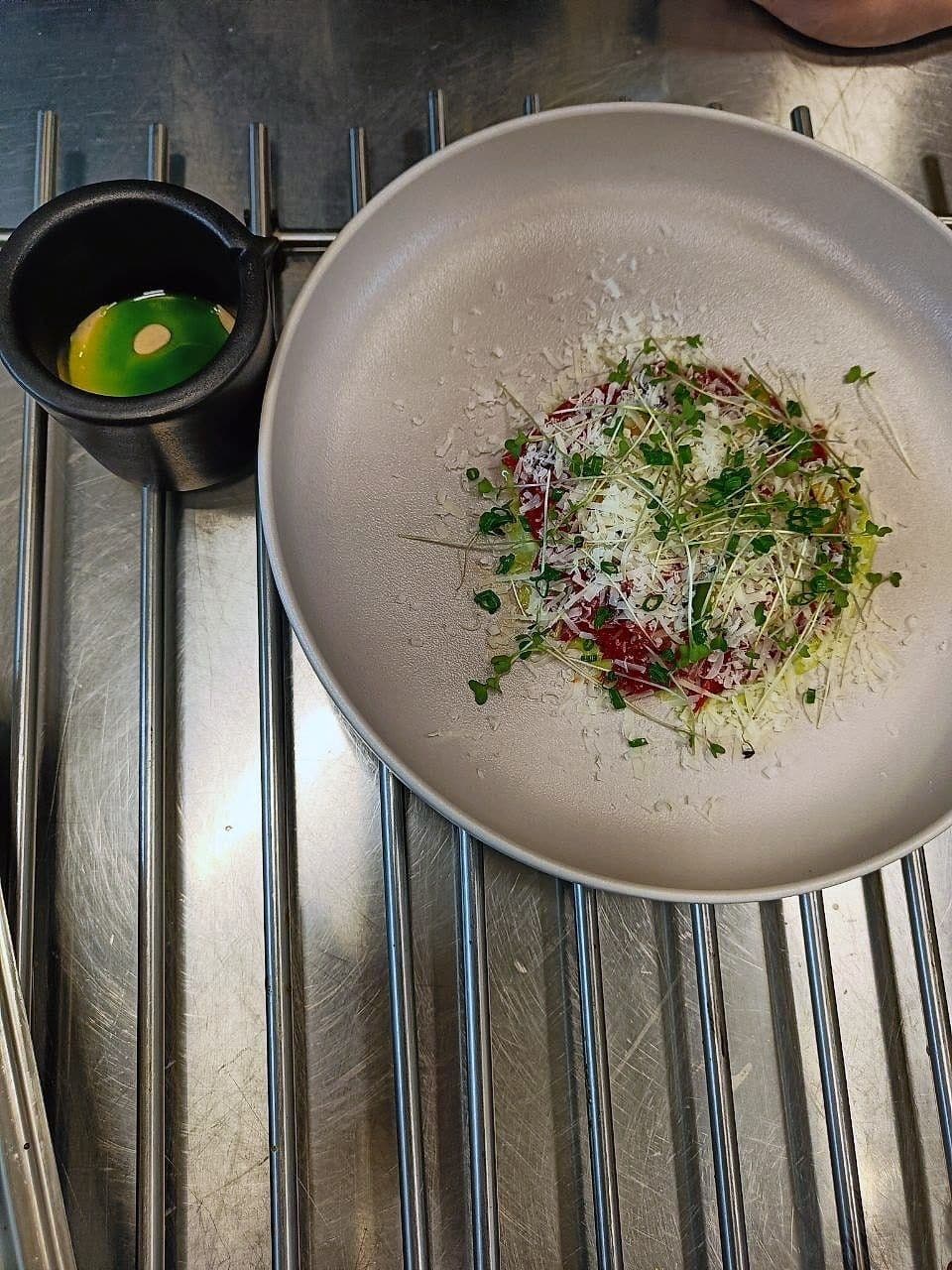 Wide disc of beef tartare covered in grated cheese and micro herbs, with a cured egg yolk in a small pot