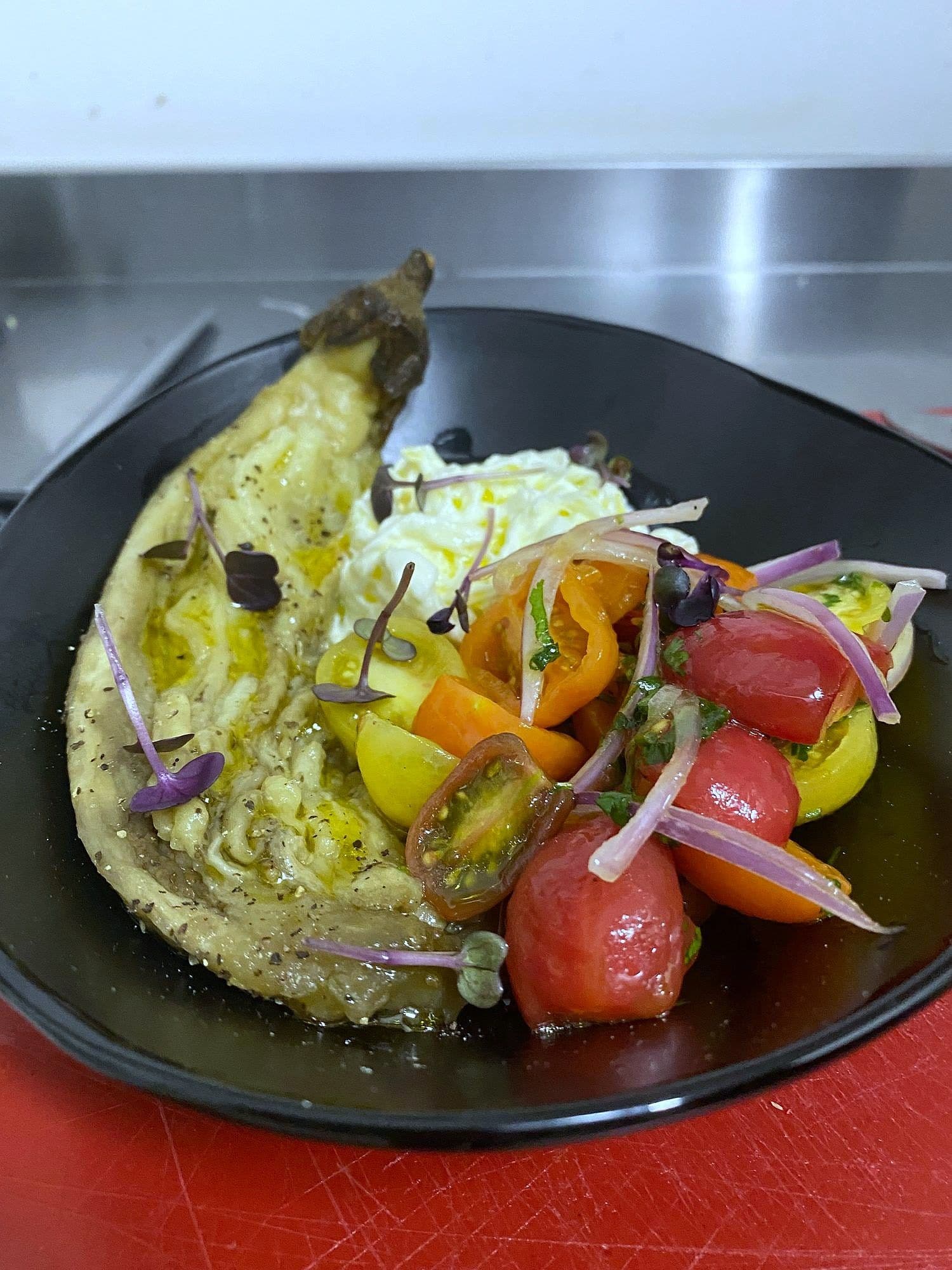 Charred aubergine split open with heritage tomatoes, pickled red onion and herbs on a black plate