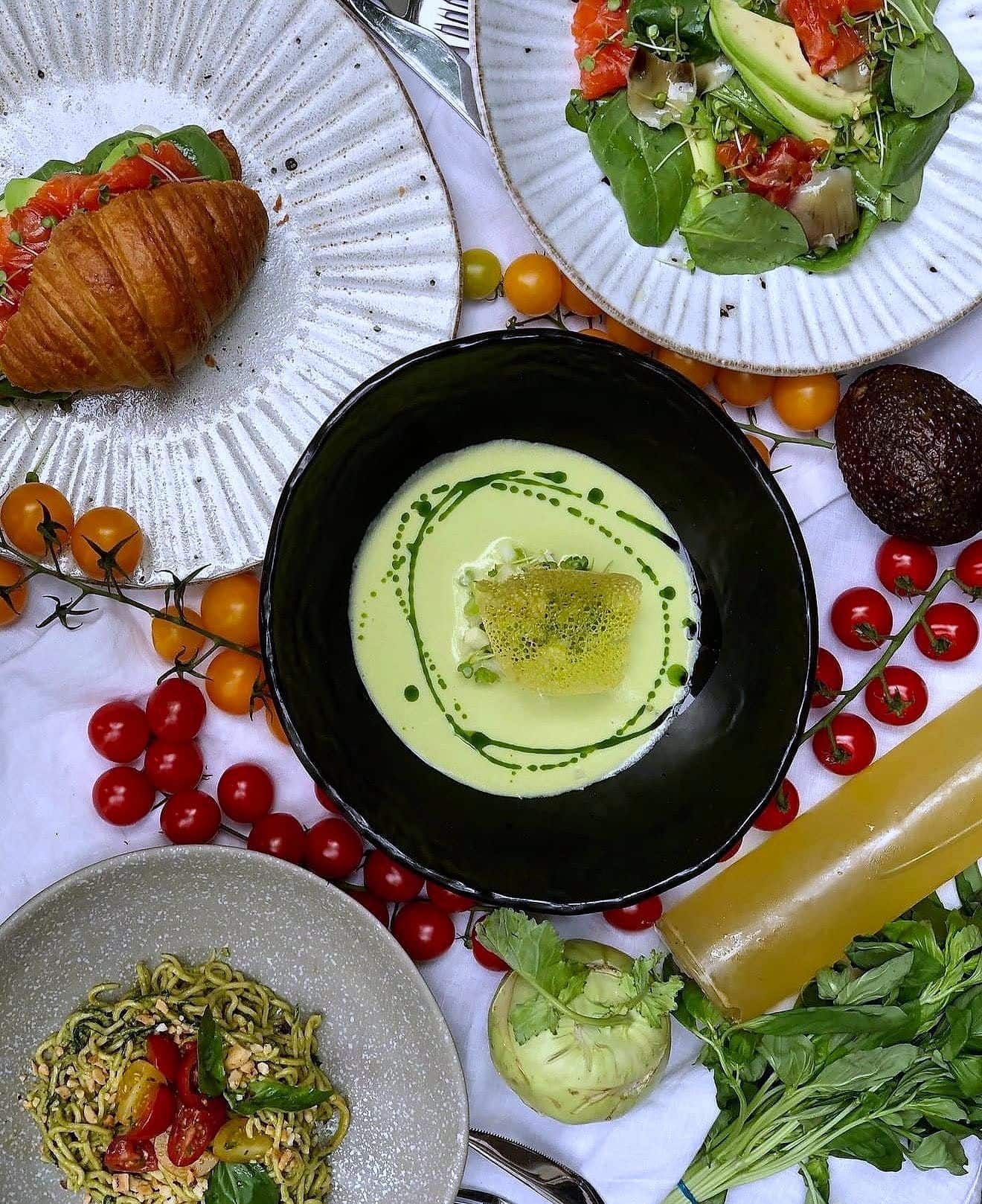 Brunch table with a filled croissant, chilled green soup, salads, cherry tomatoes and cold vegetables