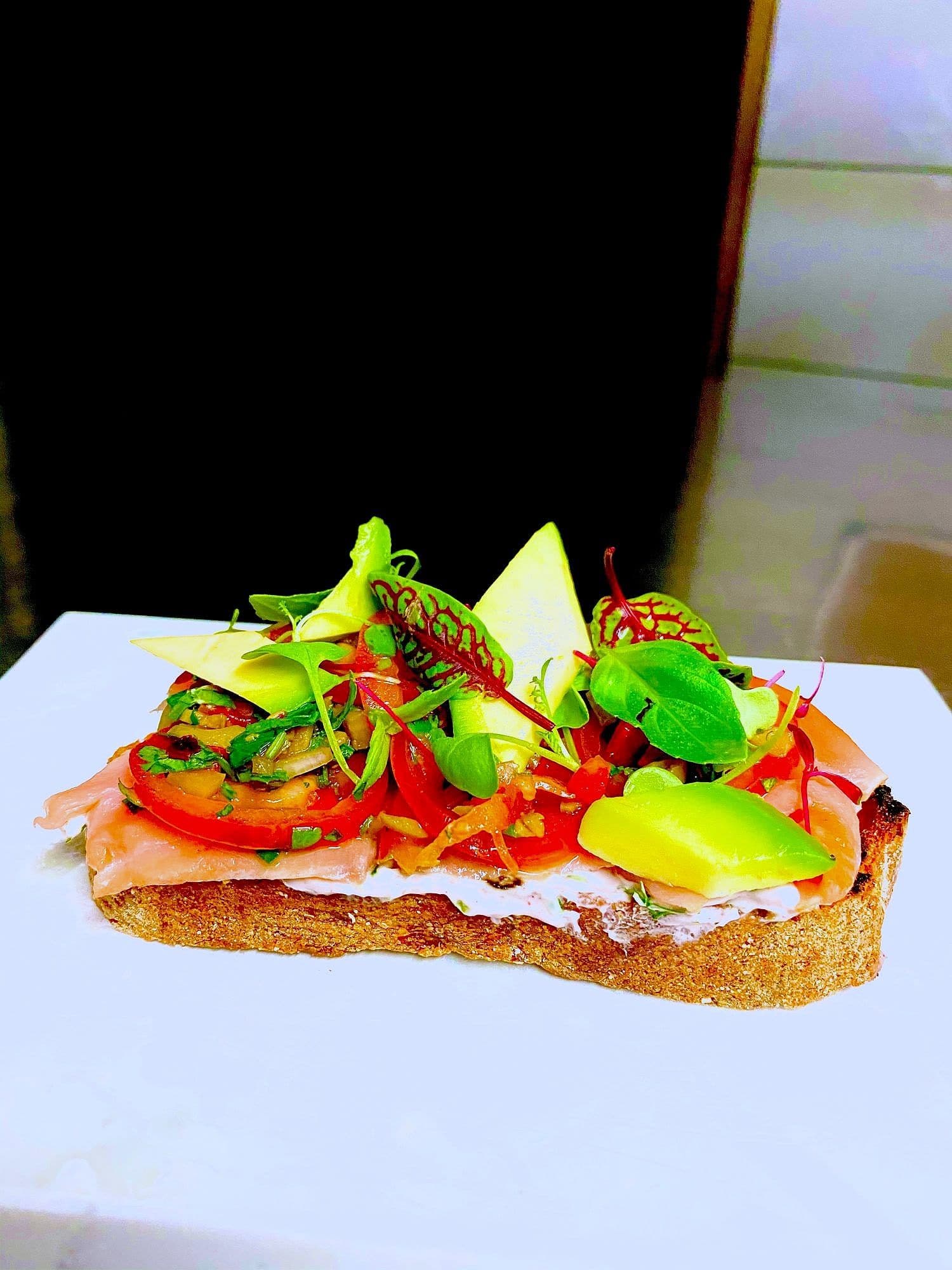 Open sandwich on sourdough with cured salmon, cream cheese, avocado, tomato and microgreens on a white board