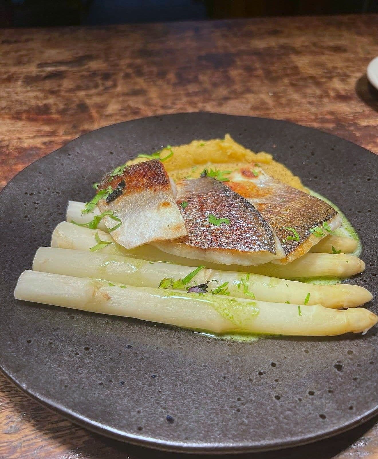 Crisp-skinned sea bass fillet over white asparagus spears with pale purée and green herb oil on a grey plate