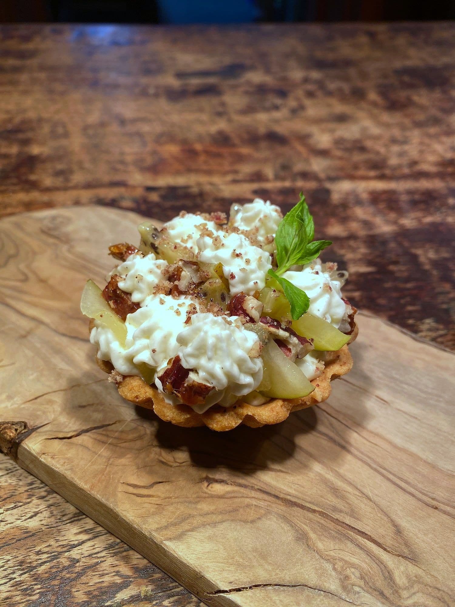 Fluted tartlet filled with piped whipped cheese, green grapes, candied pecans and a mint sprig on an olive wood board