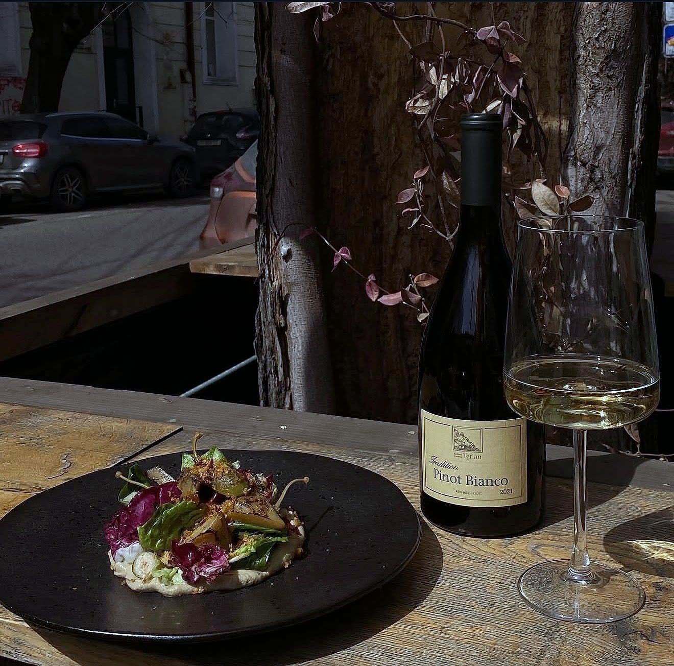 Composed grilled vegetable salad with bitter leaves on a whipped base, on a black plate beside a bottle of Pinot Bianco