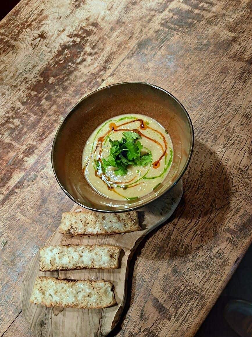 Whipped bean purée in a dark bowl drizzled with chilli and herb oil, topped with coriander, with sliced toasted bread on a wooden board