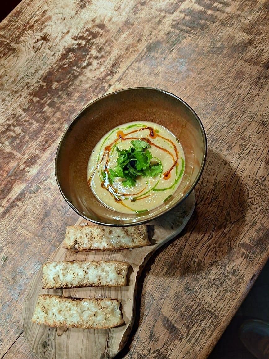 Whipped bean purée in a dark bowl drizzled with chilli and herb oil, topped with coriander, with sliced toasted bread on a wooden board