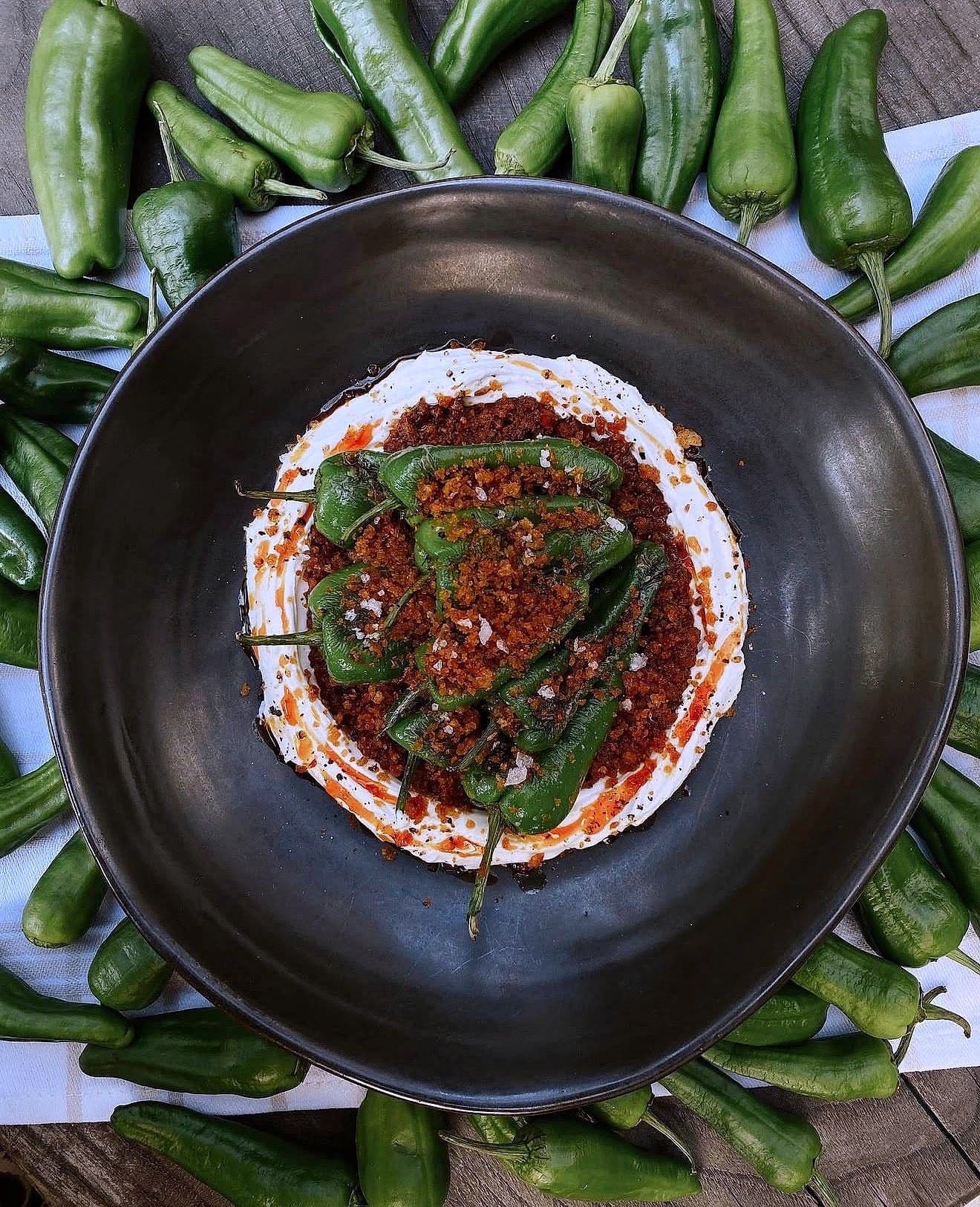 Blistered padrón peppers over labneh with a red chilli crumb in a black bowl, ringed by raw green peppers