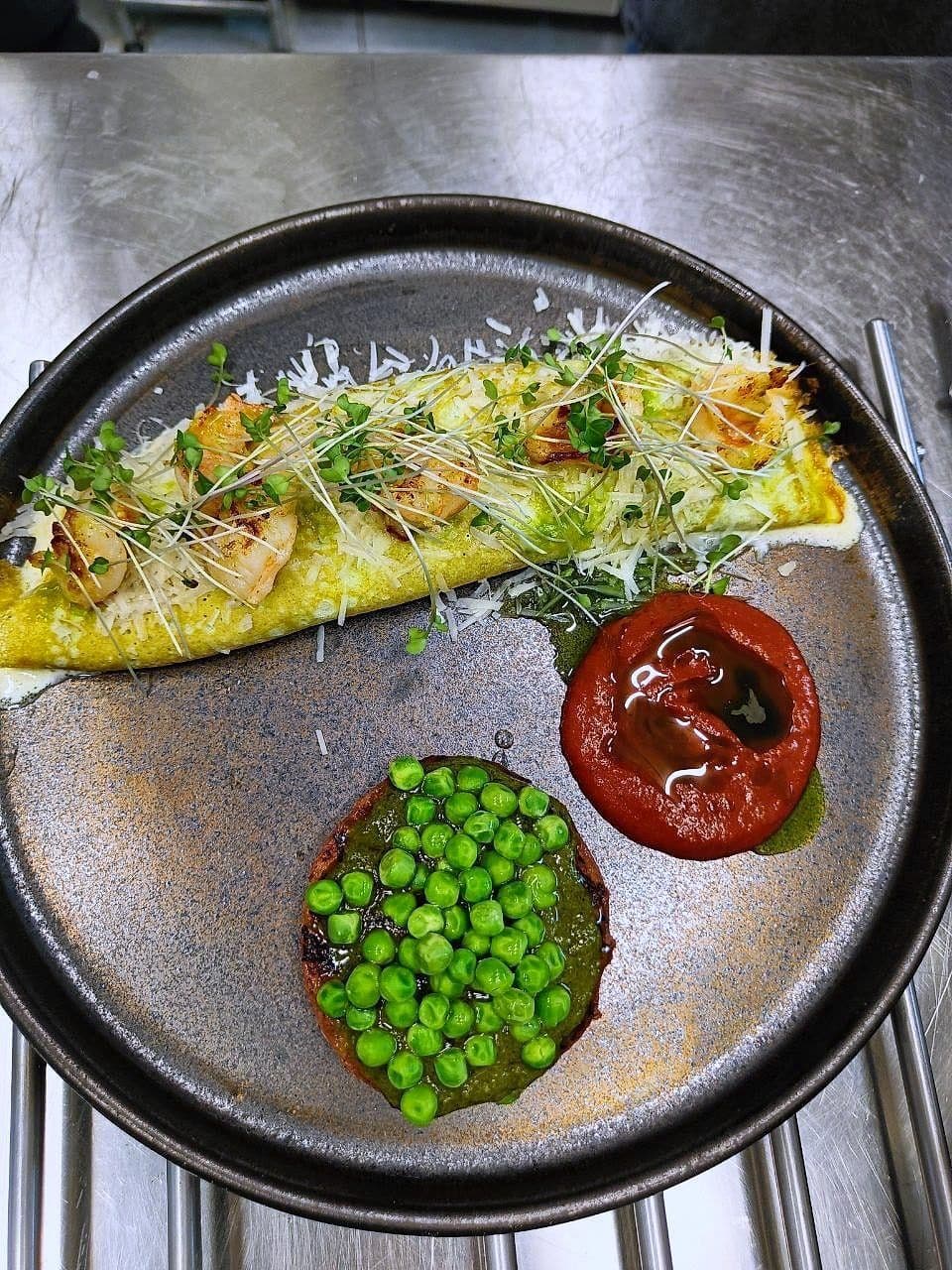 Folded omelette with cheese and micro herbs on a dark plate with peas and a slow-roast tomato