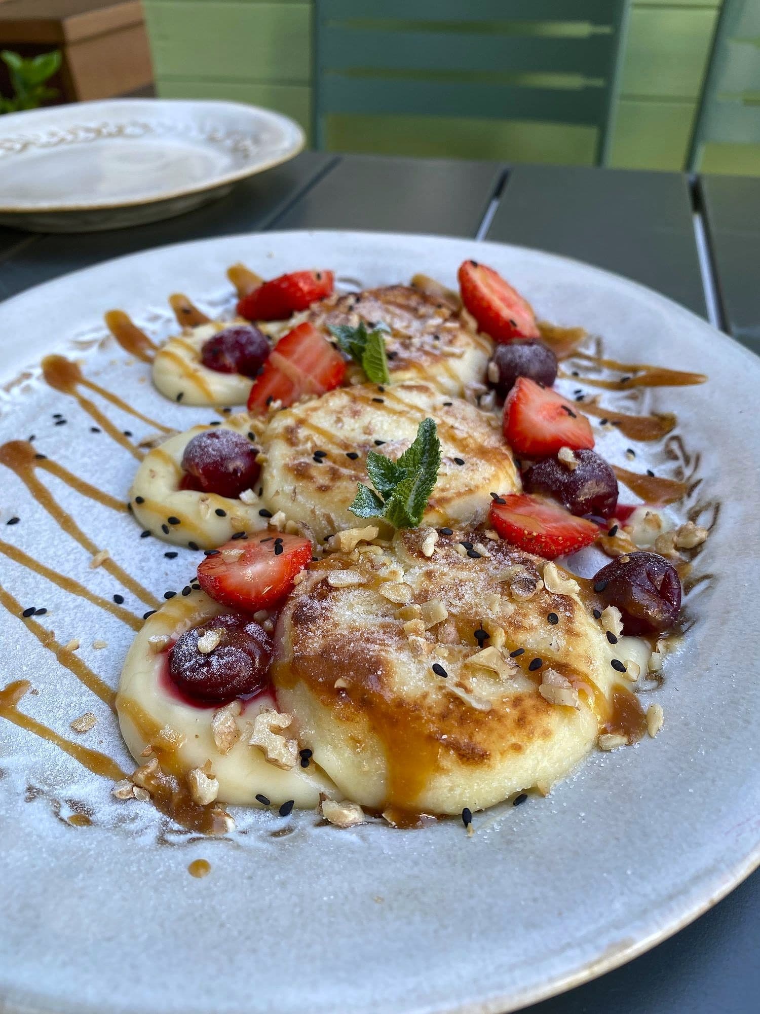 Golden syrniki cheese pancakes on a speckled plate with strawberries, cherries, caramel drizzle and sesame seeds
