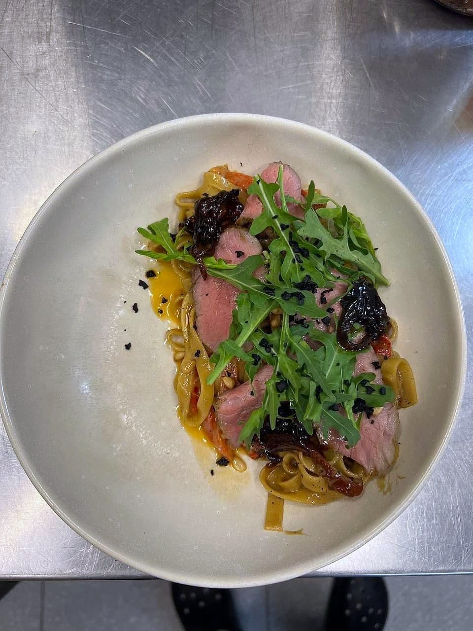 Sliced pink duck breast over pasta ribbons with rocket, dried olives and warm dressing in a pale bowl
