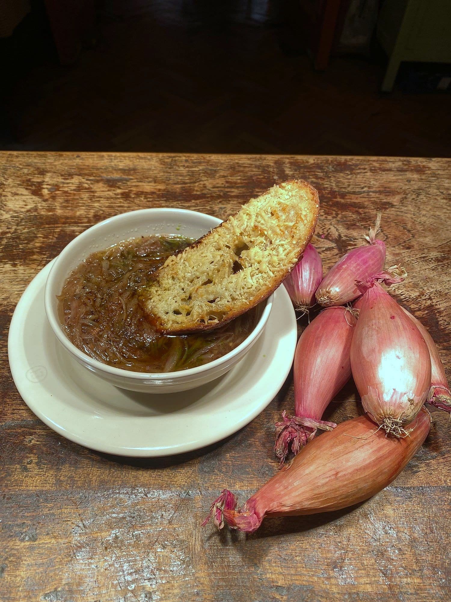 Dark onion soup in a white bowl with a slice of toasted bread on top, with fresh shallots alongside