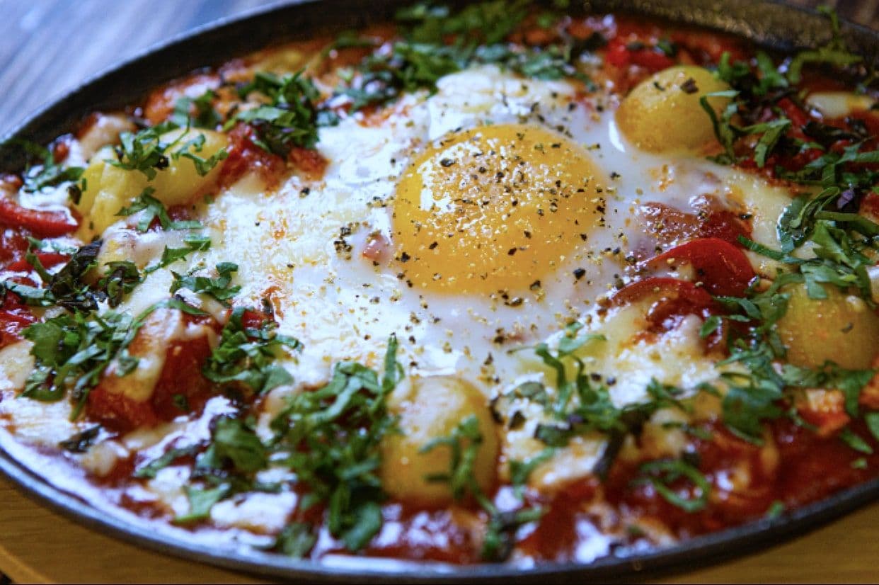 Close view of shakshuka with baked egg, tomato sauce, potato and fresh herbs in a dark pan
