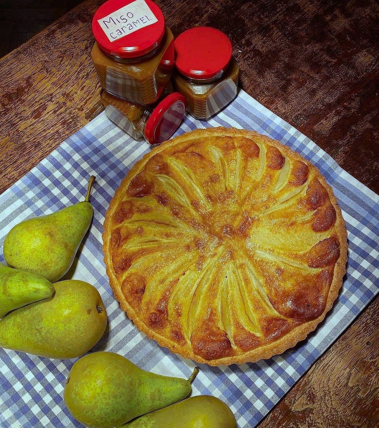Round pear tart with fanned pear slices on a checked cloth, with fresh pears and jars of miso caramel