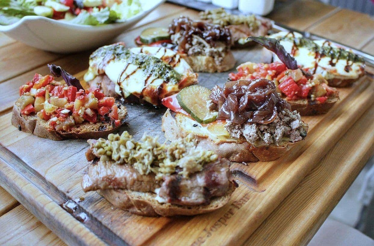 Wooden board of open bruschetta with tomato, soft cheese, cured meat and herb toppings