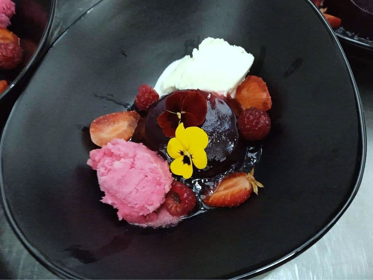 Glazed chocolate dome with raspberry sorbet, fresh berries, cream and an edible viola on a black plate