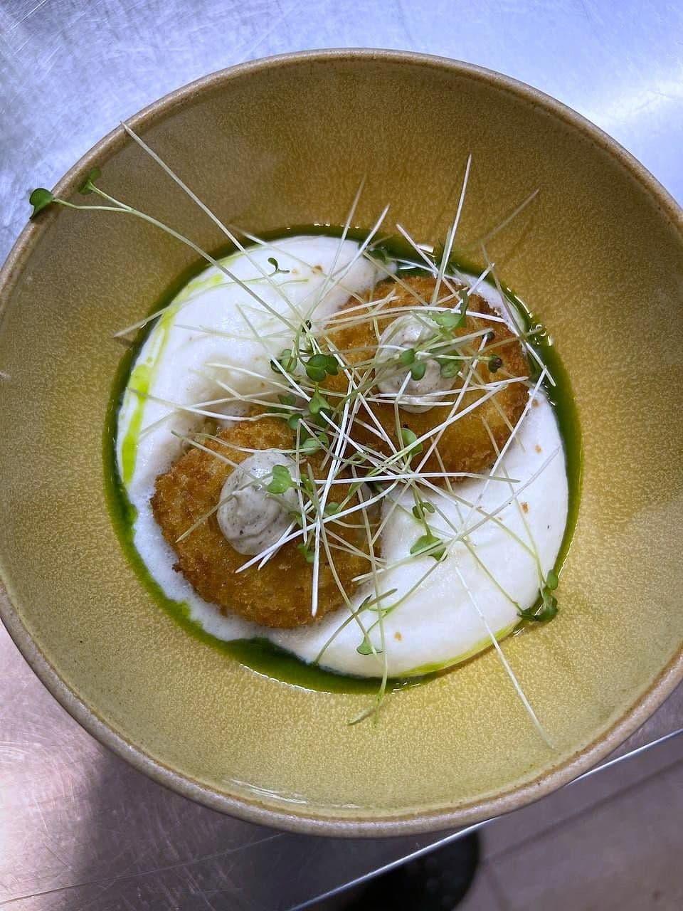 Crumbed fried cheese disc on a white base ringed with green herb oil, topped with micro herbs
