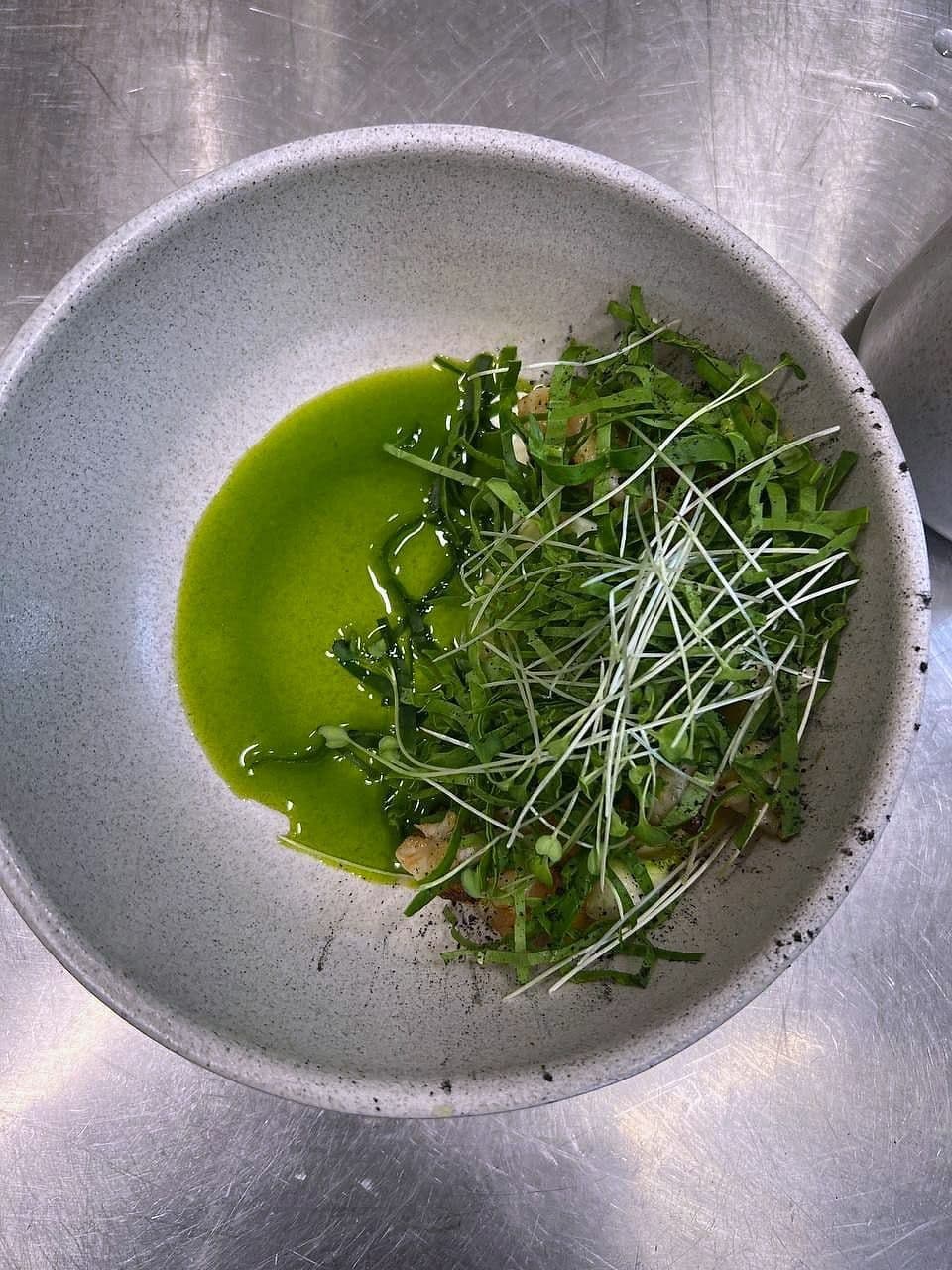 Pea shoots and cut herbs piled over bright green herb oil in a wide pale bowl