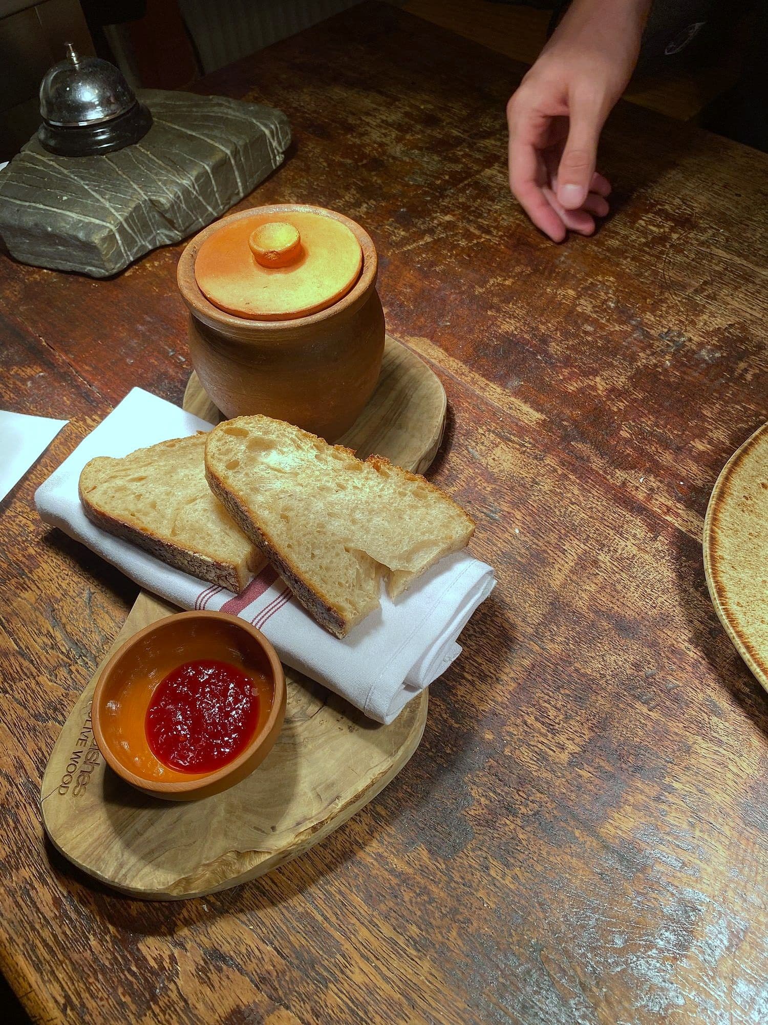 Lidded terracotta pot on a wooden board with grilled bread and a small dish of tomato sauce