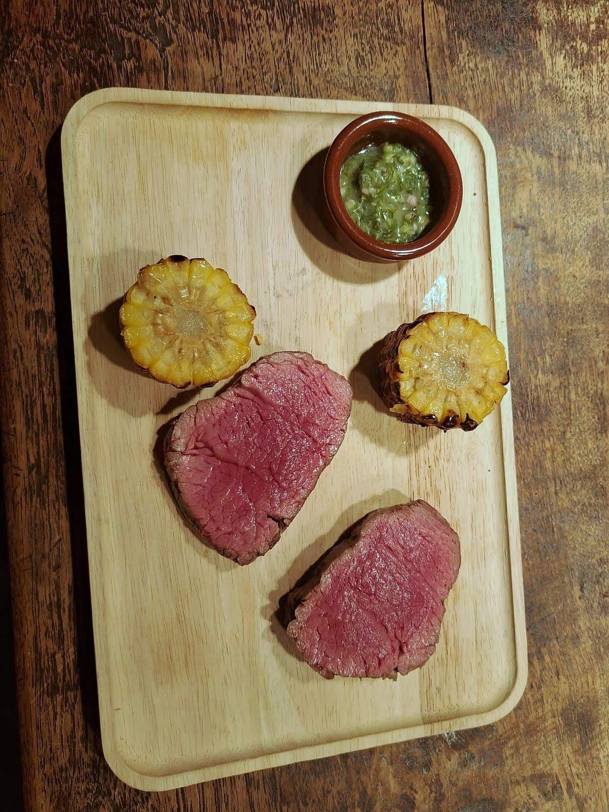 Slices of rare roast beef on a wooden board with grilled corn rounds and a pot of green herb sauce