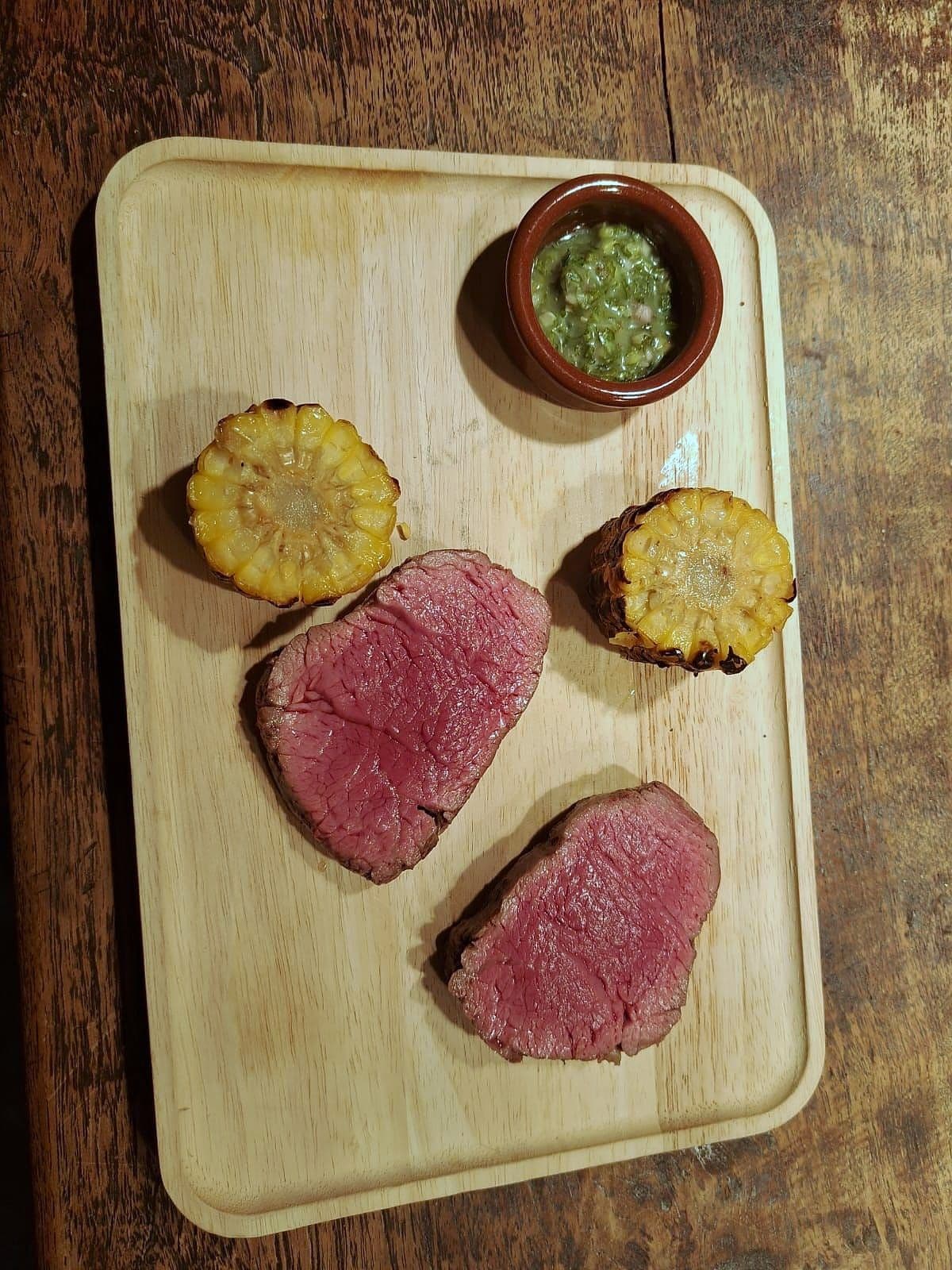 Slices of rare roast beef on a wooden board with grilled corn rounds and a pot of green herb sauce