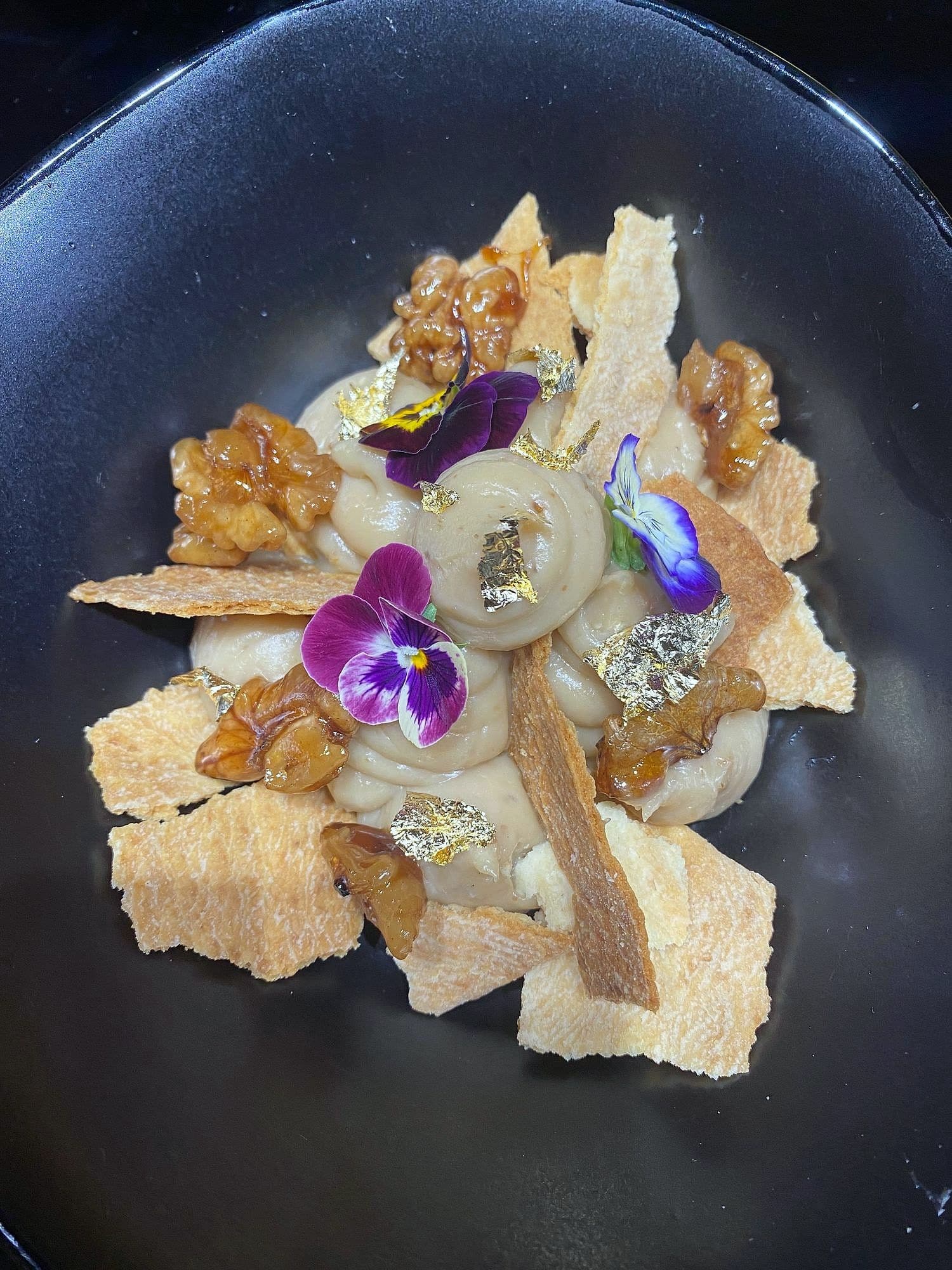 Composed plate with whipped base, candied walnuts, crisp shards, gold leaf and purple violas on dark stoneware