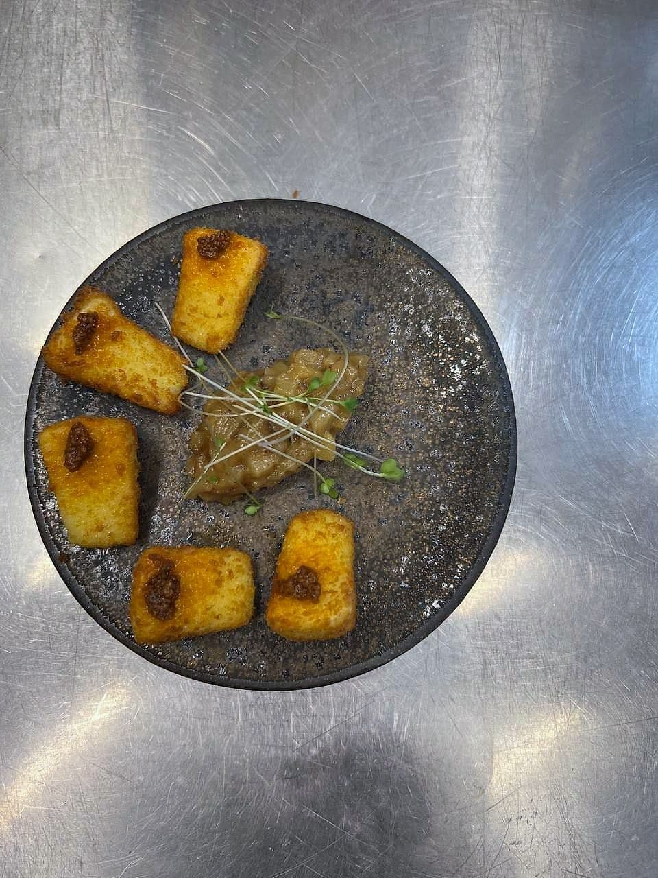Fried golden polenta cubes with confit garlic and micro herbs on a dark stoneware plate