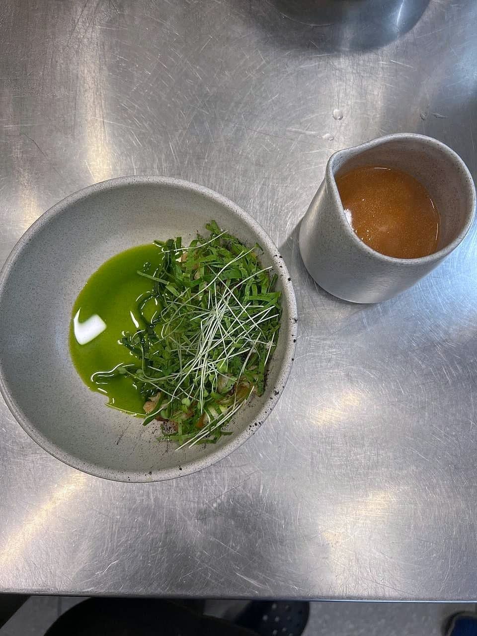 Bowl of pea shoots and herb oil beside a small jug of clear broth on a stainless bench