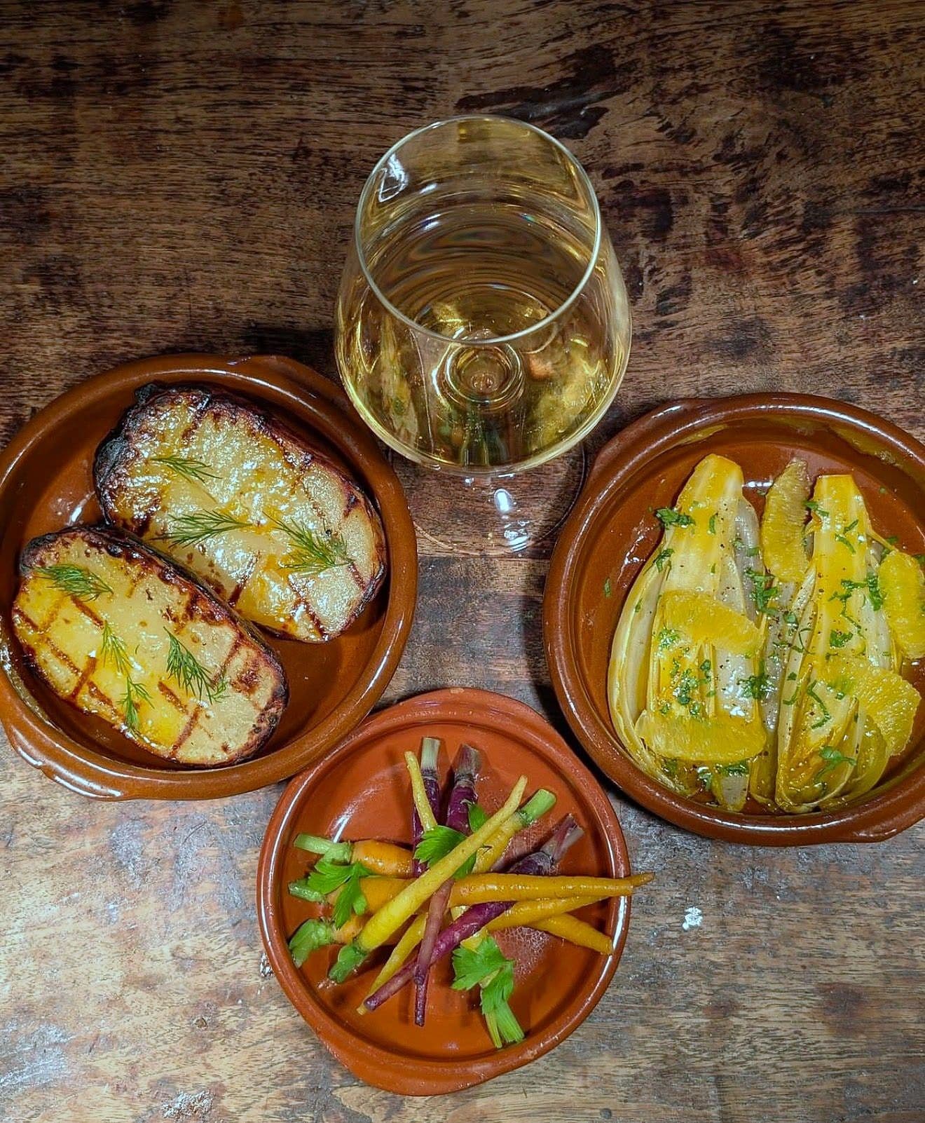 Three terracotta dishes of grilled cheese, marinated yellow peppers and pickled carrot and beetroot, with a glass of white wine