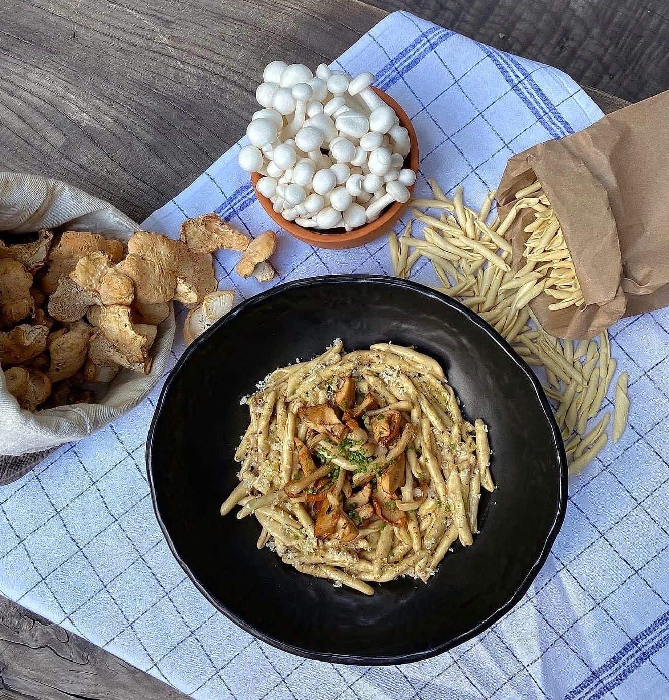 Hand-rolled short pasta with wild mushrooms in a black bowl, surrounded by raw shimeji and dried mushrooms