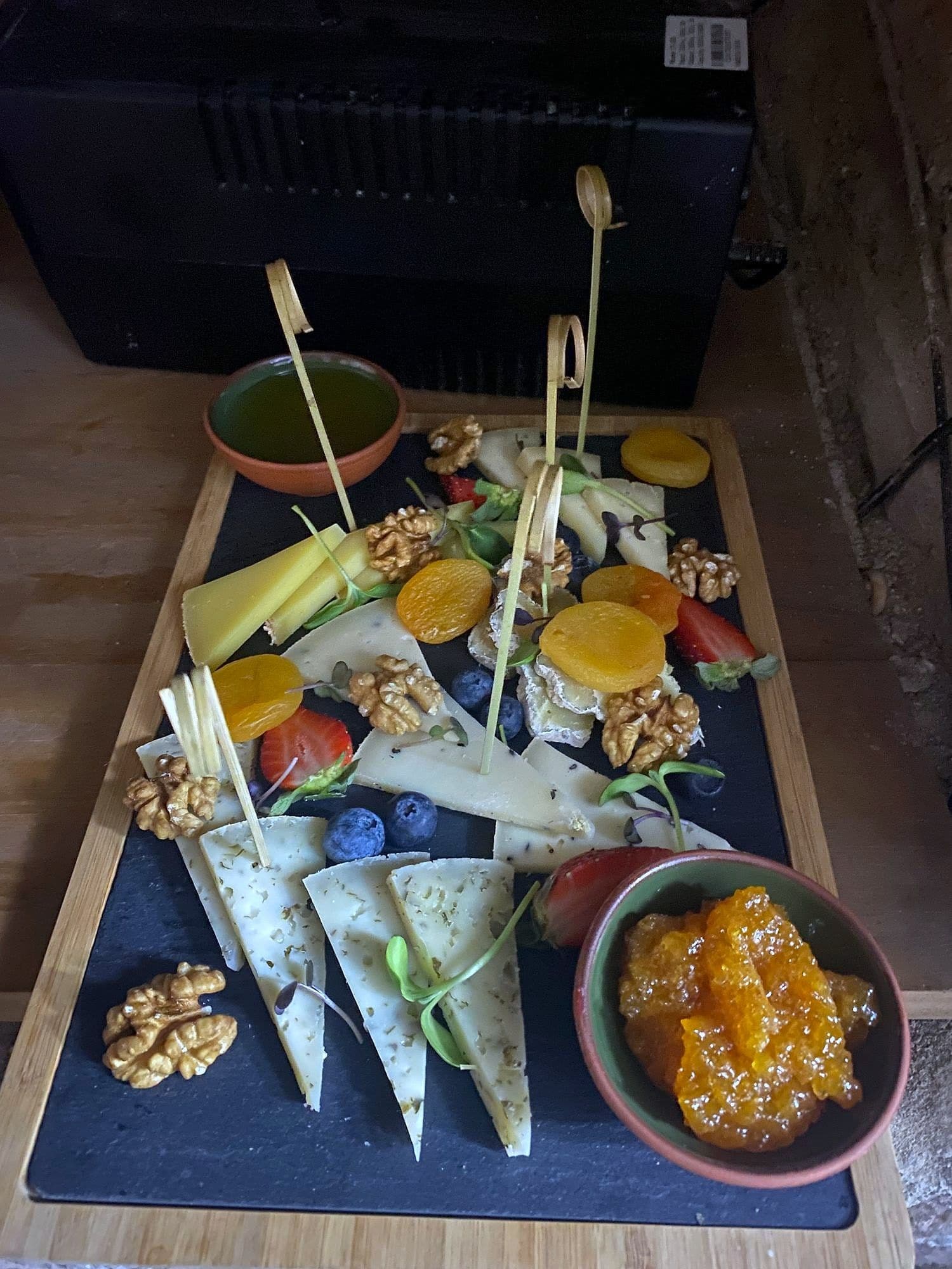 Slate board with upright wedges of cheese on skewers, walnuts, apricots, berries and a pot of fruit preserve